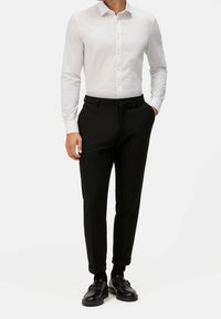 Chemise blanche boutonnée et pantalon noir, avec une coupe ajustée. Les chaussures sont noires au design élégant. Le tissu semble lisse et léger.