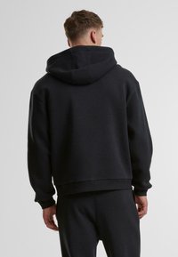 Sweatshirt noir à capuche avec une coupe décontractée, présentant une texture douce et des poignets côtelés. La vue arrière montre un design simple et sans ornement.
