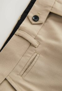 Beige Stoffhosen mit einem schwarzen Knopf, Seitentaschen und detaillierten Nähten. Nahaufnahme der weichen Textur und des strukturierten Designs.