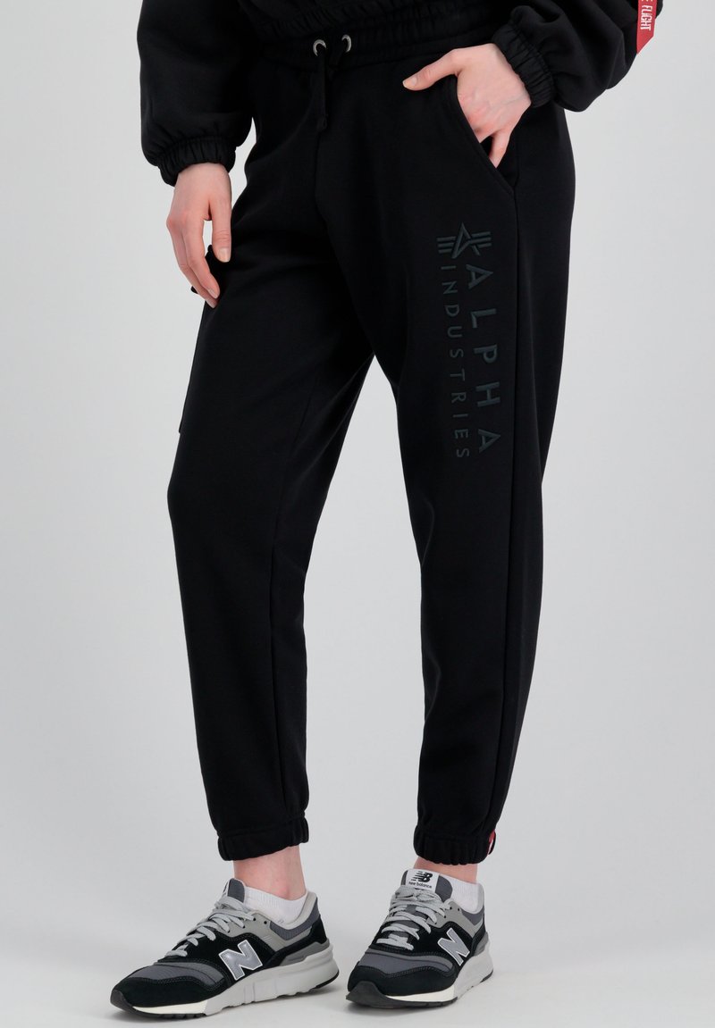 Schwarze Jogginghosen mit Seitentaschen, elastischen Bündchen, verstellbarem Bund und einem matten Logo-Print auf dem linken Bein, kombiniert mit grauen Turnschuhen.