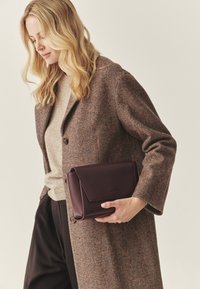 Rjava ribasta plašč čez bež pulover, v roki temnordeči usnjeni kvačkast mošnjič z zložnim zapiranjem, gladke teksture, minimalno označevanja blagovne znamke.