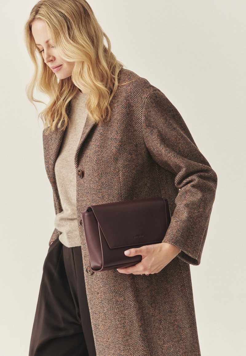 Rjava ribasta plašč čez bež pulover, v roki temnordeči usnjeni kvačkast mošnjič z zložnim zapiranjem, gladke teksture, minimalno označevanja blagovne znamke.