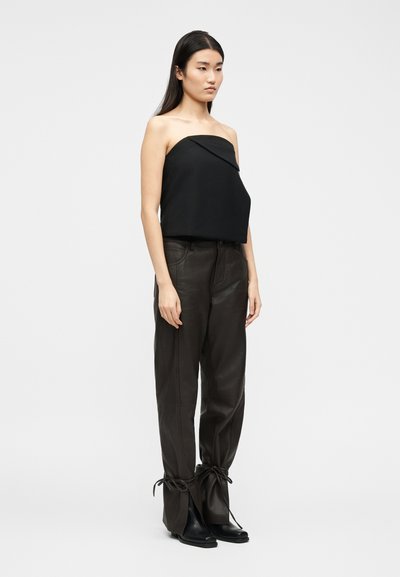 Γυναίκα που φοράει μαύρο τοπ χωρίς τιράντες, μαύρο δερμάτινο παντελόνι με δεμένα τελειώματα και μαύρες μπότες με μυτερή μύτη, που στέκεται μπροστά σε απλό λευκό φόντο.