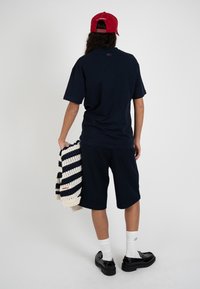 Persona vista por detrás con gorra roja, camisa y pantalones cortos azul marino, calcetines blancos, zapatos negros, sosteniendo un suéter a rayas crema y azul marino.