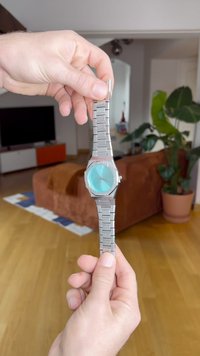 Hõbedane metallkell, millel on ümmargune türkiissinine kellasekundi, peened tunni markerid ja linkrand, hoiab käsi uduses taustas.