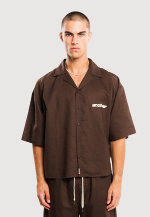 Männliches Model mit kurzen Haaren, das ein lockeres, braunes Kurzarm-Hemd mit Knöpfen trägt, auf dessen Brust das Wort "another" steht, sowie passende Hosen mit weißen Kordelzügen.