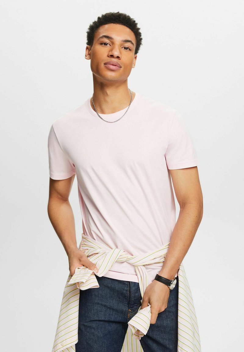 Helles rosa T-Shirt mit kurzen Ärmeln aus glattem Stoff, kombiniert mit dunklen Jeans und einem um die Taille gebundenen, weiß gestreiften Hemd. Silberne Kettenhalskette.