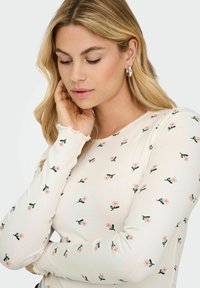 Langærmet top i cremefarvet ribbet stof med grønt blomstermønster. Har rund halsudskæring og bølgede manchetter.