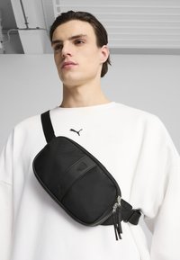 Valigia a tracolla nera con rete testurizzata e dettagli in pelle liscia. Presenta una chiusura con zip e una tracolla regolabile. Indossata diagonalmente sul corpo.