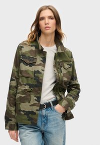 Camo-jack in groene en zwarte tinten, met knoopzakken op de borst, een kraag en een ritssluiting vooraan, gecombineerd met een grijs shirt en blauwe spijkerbroek.