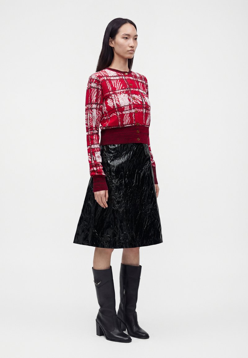 Femme portant un cardigan rouge à motifs, une jupe noire brillante longueur genou et des bottes noires à talons hautes jusqu'aux genoux, debout devant un fond uni.
