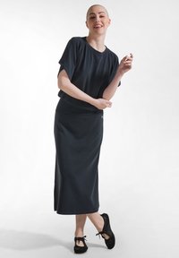 Vrouw in een zwart T-shirt met korte mouwen, zwarte midi-rok en zwarte veterschoenen, staand met één voet gekruist, glimlachend tegen een witte achtergrond.