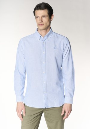 Uomo con camicia a righe azzurro chiaro e bianca con bottoni e pantaloni verde oliva in piedi davanti a uno sfondo semplice.