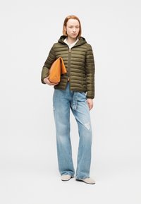 Γυναίκα με μπουφάν σε ελιά πράσινο, ανοιχτό μπλε τζιν με φαρδιά, σκισμένα μπατζάκια, μπεζ παπούτσια, κρατώντας μεγάλη πορτοκαλί τσάντα φάκελο, στέκεται μπροστά σε λευκό φόντο.