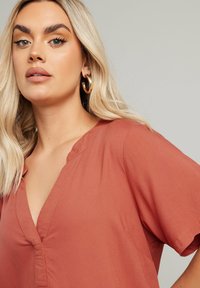 Rostfärgad blus med V-ringning och korta ärmar, med en slät textur och subtila sömnadsdetaljer. Guldörhängen i form av hoops synliga.