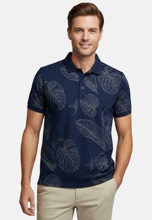 Man in een marineblauw poloshirt met grote bladerenprints en beige broek, staand met één hand in de zak.