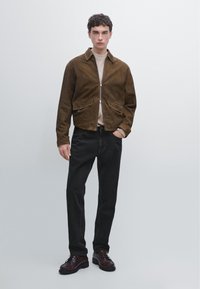 Brun ruskindsjakke med lynlås foran, to påsyede lommer, båret over en lys beige sweater, sammen med sorte jeans og mørkebrune sko.