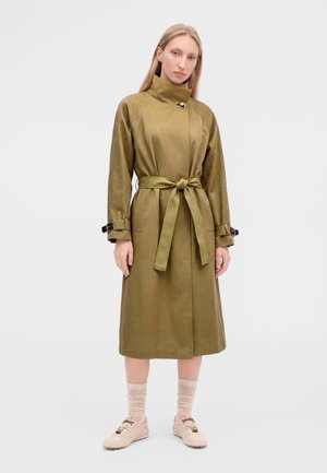 Femme portant un long manteau ceinturé vert olive avec un col montant, debout les mains détendues sur les côtés, chaussettes claires et baskets beiges.