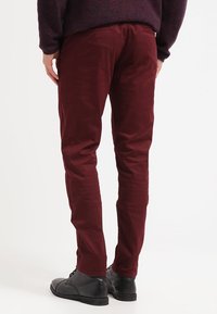 Pantaloni in velluto a coste color bordeaux con vestibilità slim, caratterizzati da una sottile trama a coste e tasche posteriori. Indossati con scarpe in pelle nere.