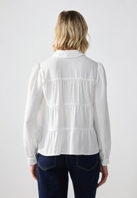 Hvid langærmet bluse med samlet detaljering, krave og knapbeslåede manchetter, parret med mørke denimjeans. Glat, letvægtsstof.