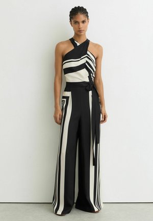 Mujer con cabello trenzado, vestida con un mono tipo halter de rayas negras y blancas, con piernas anchas y cinturón en la cintura, de pie frente a una pared lisa.