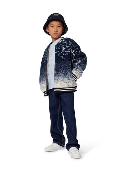 Blouson bomber bleu et blanc à motifs nuages, t-shirt graphique bleu clair, pantalon en denim foncé, chaussures blanches et chapeau seau bleu marine.