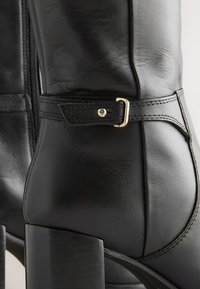 Bottine en cuir noir avec une silhouette courbée, une fermeture éclair latérale et une bride décorative avec une boucle dorée. Talon épais.