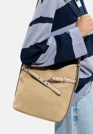 Osoba w niebiesko-szarym paskowanym swetrze trzyma beżową skórzaną torbę na ramię z regulowanym paskiem i srebrnymi okuciami.