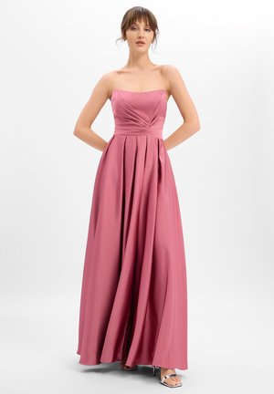 Frau in einem trägerlosen, staubrosafarbenen Satinkleid mit plissiertem Oberteil und fließendem Rock, die mit den Händen hinter dem Rücken vor einem weißen Hintergrund steht.