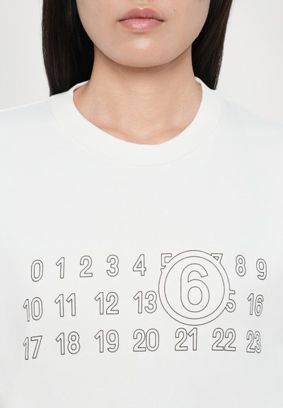 Felpa bianca con numeri neri disposti a griglia, con un design circolare prominente attorno al numero 6 al centro.