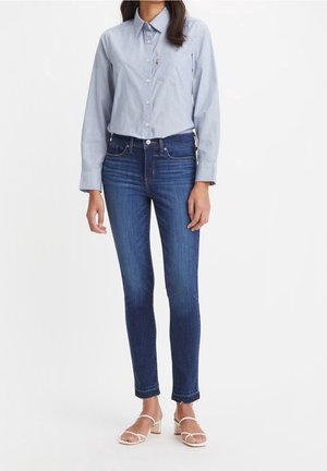 Camisa azul de botones con cuello y bolsillo, combinada con jeans skinny de mezclilla oscura y sandalias claras; texturas suaves de algodón y mezclilla.