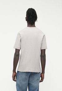 Tricou gri din bumbac cu mânecă scurtă, decolteu rotund, suprafață texturată și croială lejeră, văzut din spate. Blugi din denim purtați sub tricou.