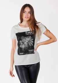 Camiseta blanca con un gráfico de palmera negra, que presenta el texto "IBIZA PART PARADIS" en negrita. Cuello redondo, mangas cortas, ajuste relajado.