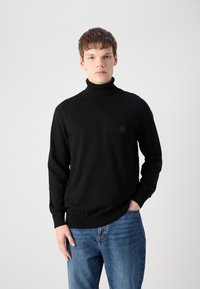 Črn pulover s turtleneck ovratnikom iz mehkega materiala, z dolgimi rokavi, prileganim ovratnikom in majhnim logotipom na prsih. Obut z modrimi kavbojkami.
