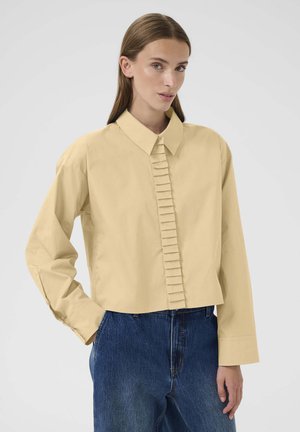 Camicia giallo chiaro tagliata, con dettagli plissettati sul davanti, colletto a punta e maniche lunghe; abbinata a jeans in denim blu.