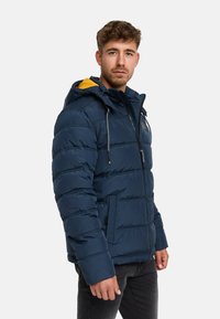 Giacca puffer blu navy con fodera della cappuccio gialla, motivo a righe orizzontali, zip frontale, stringhe di regolazione e tasche laterali.