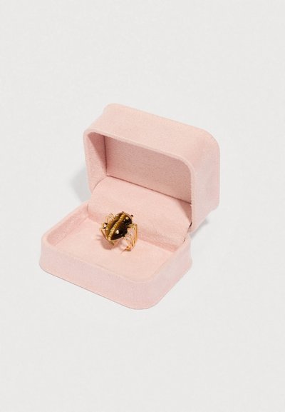 Kuldne ja must krabi kuju sõrmus, mis on välja pandud pehmes roosas ehtekarbis, mille sees on pehme polsterdus; karbil on siledad servad ja ümarad nurgad.