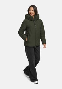 Woman wearing a dark green puffer jacket with high collar, black wide-leg pants, and white sneakers, smiling and walking with one hand in pocket.