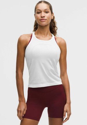 Junge Frau trägt weißes ärmelloses Oberteil und burgunderrote Sportshorts, steht mit entspannten Händen vor einfachem Hintergrund.