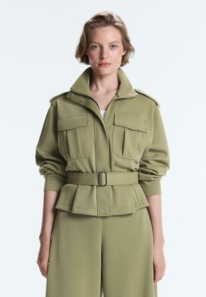 Jeune femme portant une veste ceinturée vert olive avec deux grandes poches poitrine et un pantalon assorti, posant devant un fond blanc uni.