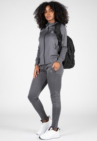 Grijze sportieve trackjack met een rits aan de voorkant, passend joggingsbroek en witte sneakers. Rugzak met zwarte mesh accenten wordt gedragen.