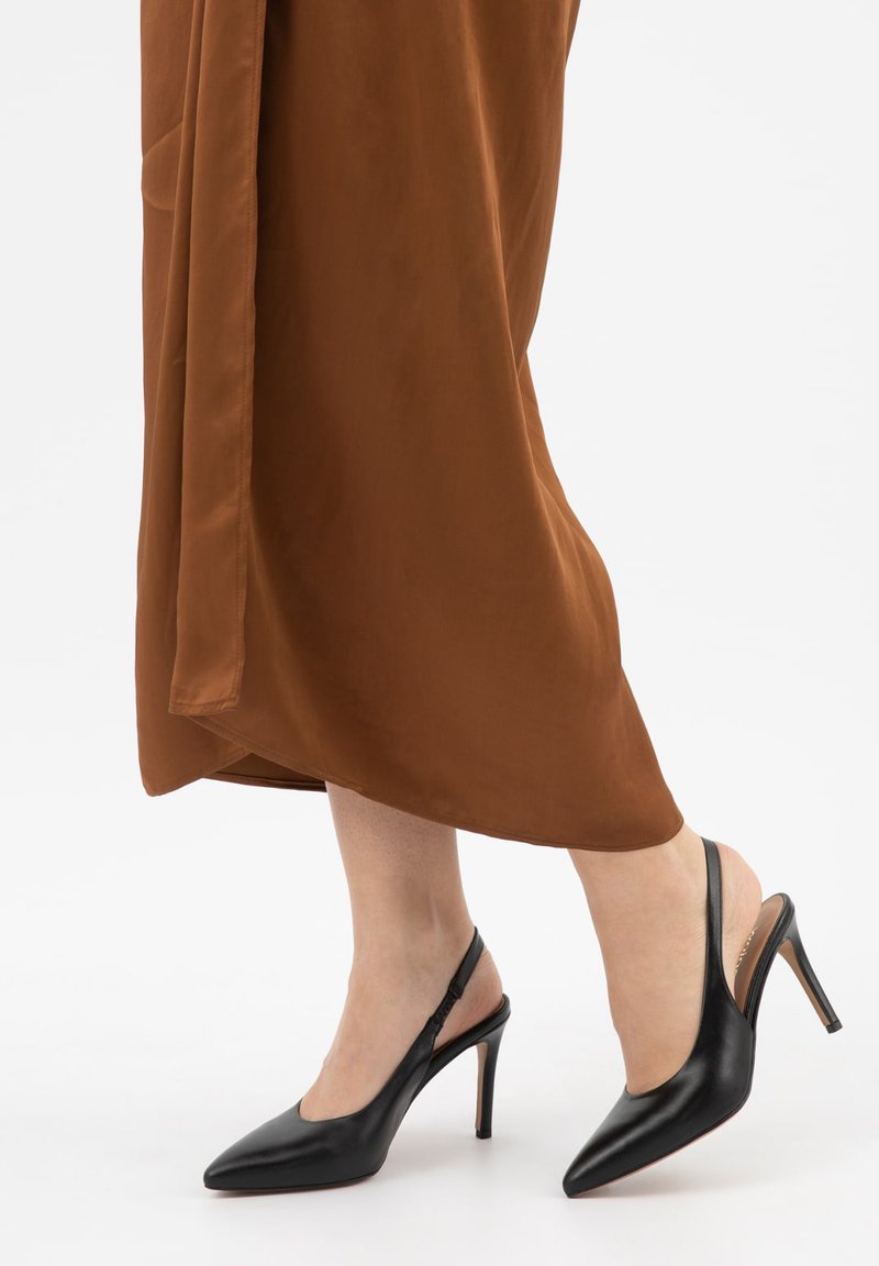 Benen met zwarte slingback hoge hakken met puntige neus, gecombineerd met een bruine midi-rok tegen een effen lichte achtergrond.