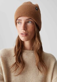 Brown ribbed knit beanie made from soft material, featuring a black tag accent. Model wears a beige sweater, with long wavy hair visible.