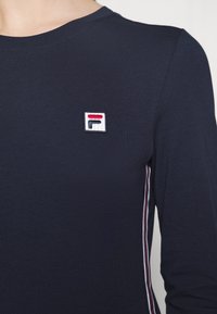 Mörkblå långärmad skjorta i mjukt bomull, med en liten broderad logotyp och kontrasterande vita och röda sidostråk.