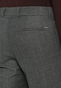 Prim-plan al unor pantaloni gri texturați, purtați cu un top închis la culoare, de culoare maro, băgat în pantaloni, care arată buzunarul din spate și mânerele pentru curea.