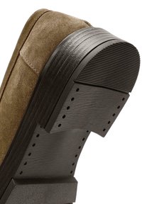 Talons de chaussures en daim marron avec semelle en caoutchouc noire texturée et coutures visibles sur la tige et les bords de la semelle.