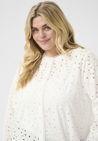 Blusa blanca con un patrón de ojales texturizados, cuello redondo y tapeta decorativa de botones. Mangas largas con hombros ligeramente abullonados.