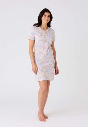 Vrouw staat blootsvoets en draagt een korte mouwen, knielange jurk met een kleurrijk bloem- en bladmoten op witte achtergrond.