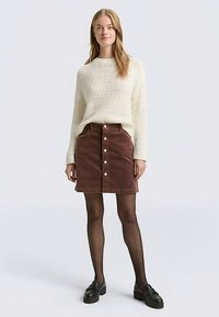 Lichtbeige gebreide trui met een gestructureerd patroon, gecombineerd met een bruine ribfluwelen rok met knopen aan de voorkant en zakken, gedragen met zwarte loafers.