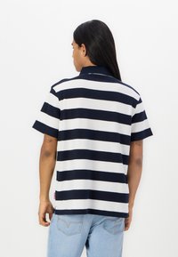 Camisa polo riscas azul-marinho e branca com mangas curtas, colarinho, tecido texturizado e fendas laterais. Usada com jeans de ganga azul-claro.